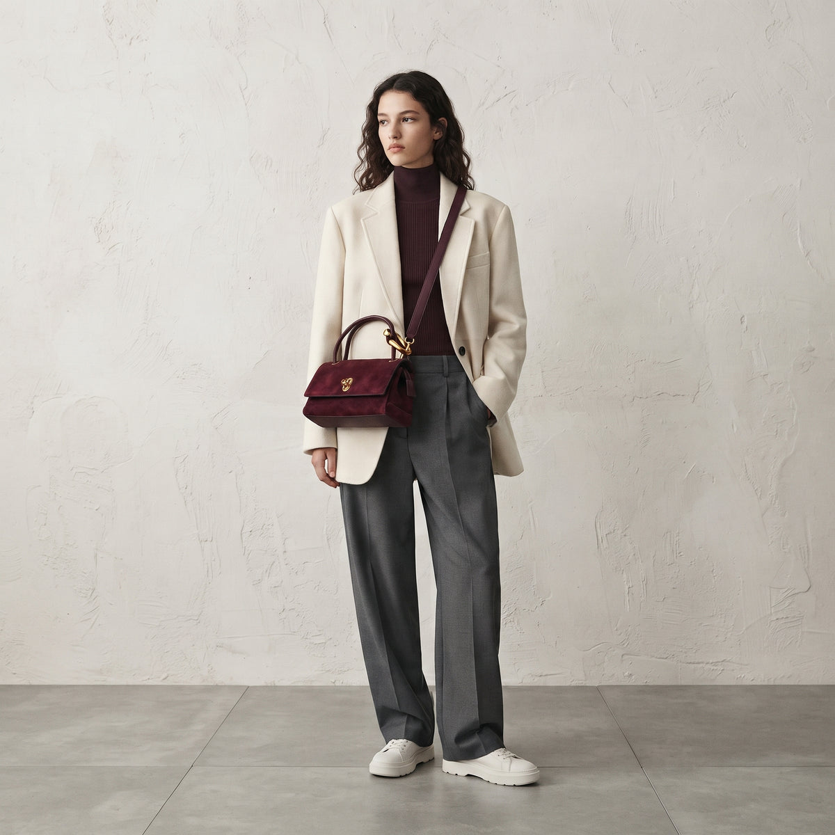 Modelo luciendo el bolso de ante modelo Elara en color burdeos con correa cruzada, combinado con un blazer blanco y pantalón gris.