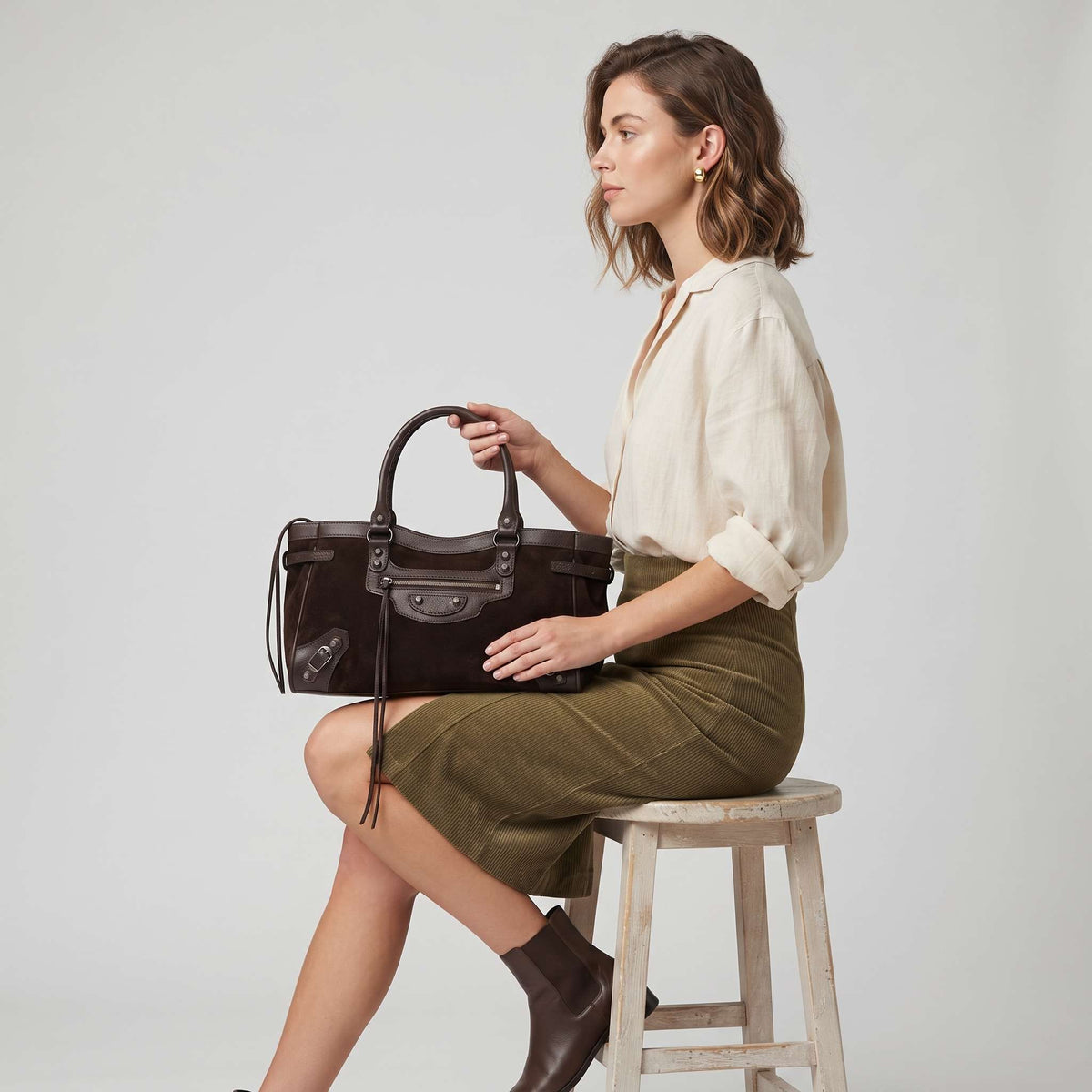 Texto alternativo (Alt Text):
Modelo sentada en un taburete sosteniendo el bolso satchel marrón modelo Cleo con detalles de cremalleras, hebillas y diseño de ante.