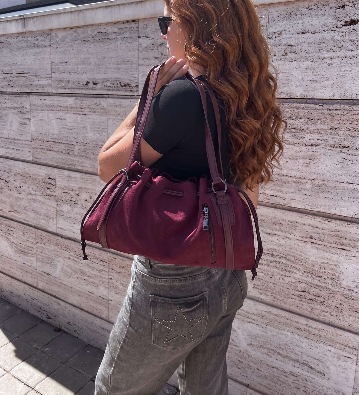 ista posterior de una modelo sosteniendo un gran bolso de hombro de serraje color burdeos de diseño fruncido. El bolso cuenta con cordones laterales, borlas y herrajes plateados. La modelo lleva una camiseta negra, vaqueros grises y gafas de sol, mostrando cómo queda el bolso puesto