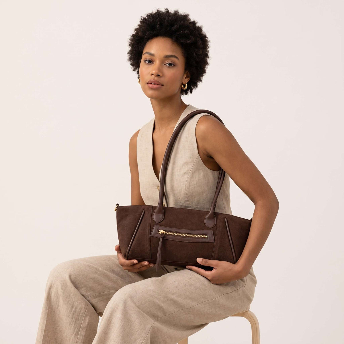 Fotografía de estudio de una mujer de raza negra con cabello afro y un traje de lino beige (chaleco y pantalones anchos) sentada y sosteniendo un bolso de hombro de ante marrón oscuro en su regazo. El bolso tiene ribetes verticales de cuero, herrajes dorados y un bolsillo central con cremallera.